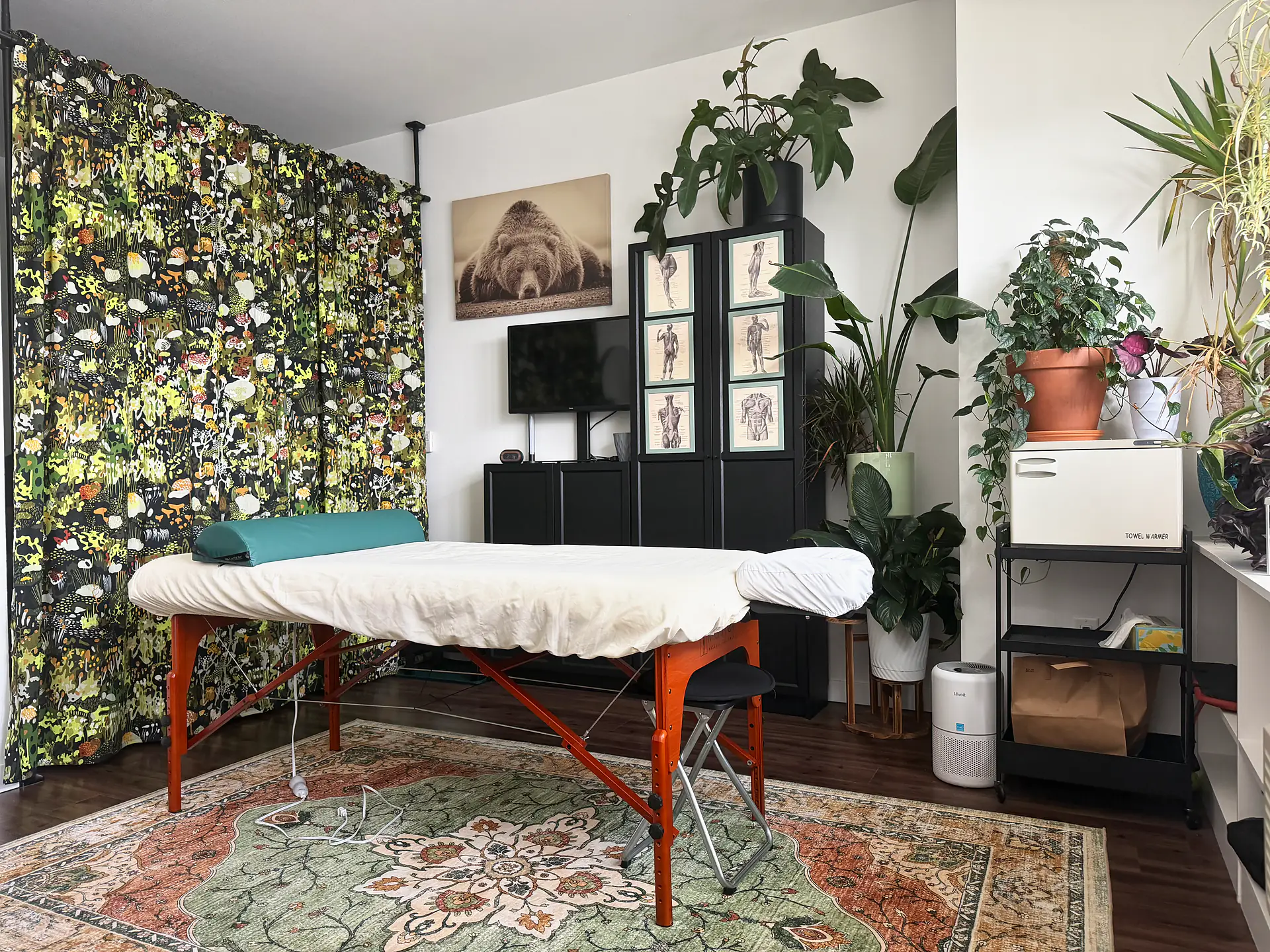 Massage table in the Portland studio below a bear print, with a woodland-pattern curtain and tall houseplants